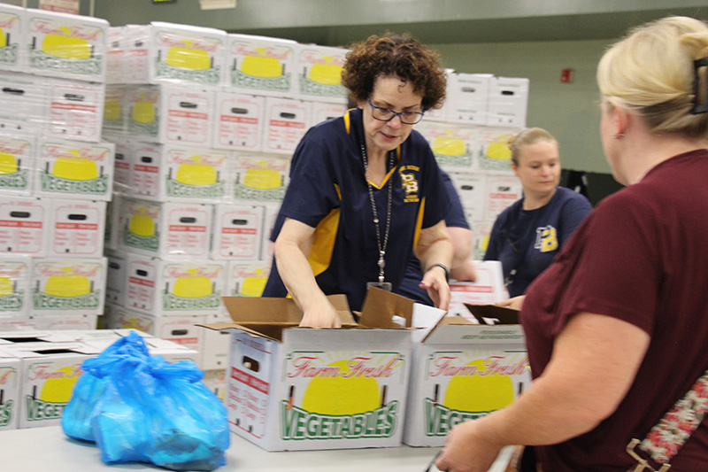 Stacks of large white and yellow produce boxes are behind  two women dressed in blue and gold who are handing out the boxes.