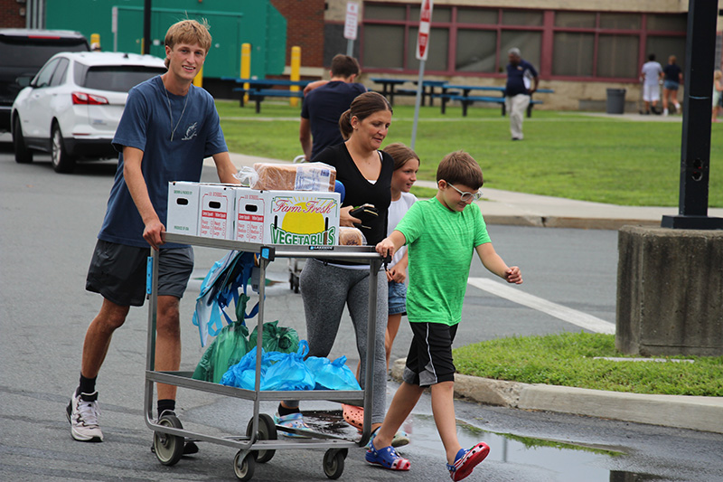 A high school boy smiles as he pushes a car with boxes of produce on it and blue bags on the bottom of the cart. There is a woman and two boys walking with him.