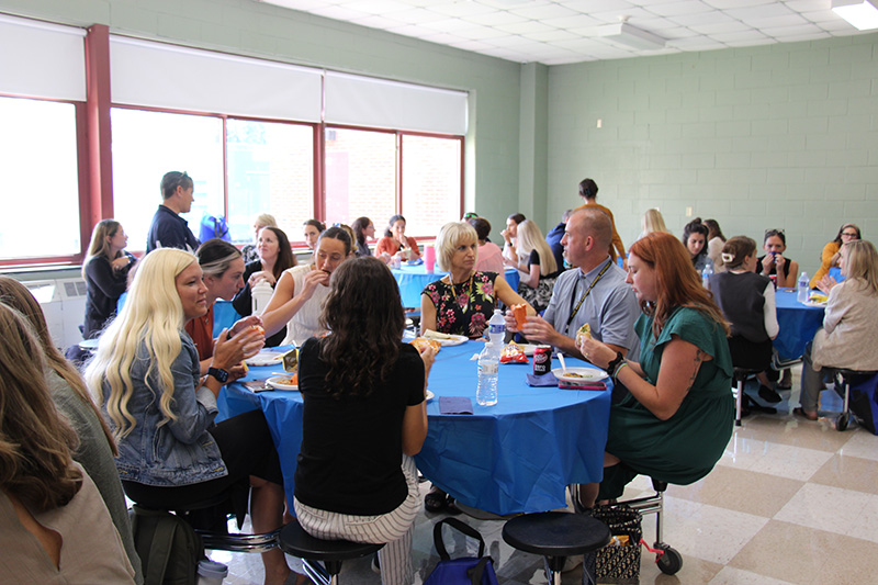 A group of adults sit around table with blue tablecloths on them. The people are eating and talking.