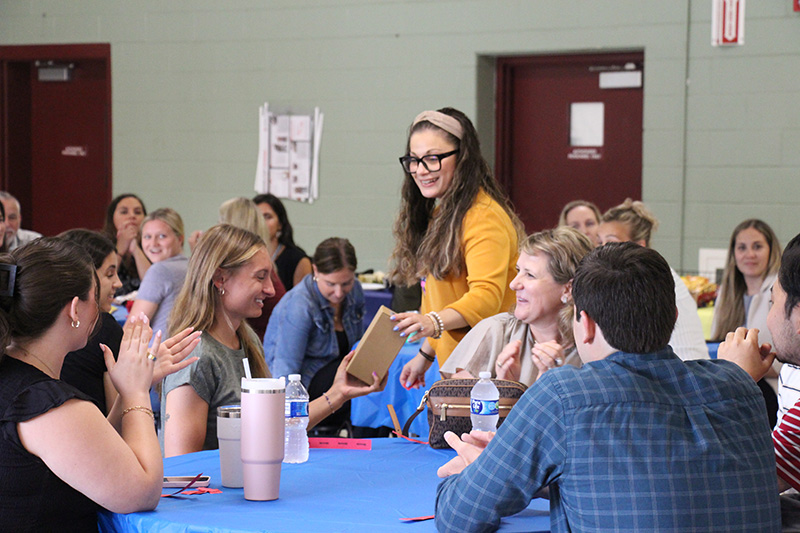 A woman with glasses, long hair and a yellow sweater hands a box to a woman sitting at a table. There are many others at the table smiling.