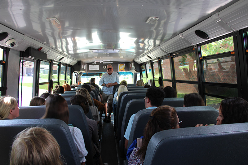 From the back of a school bus, a man talks to adults sitting on the bus.