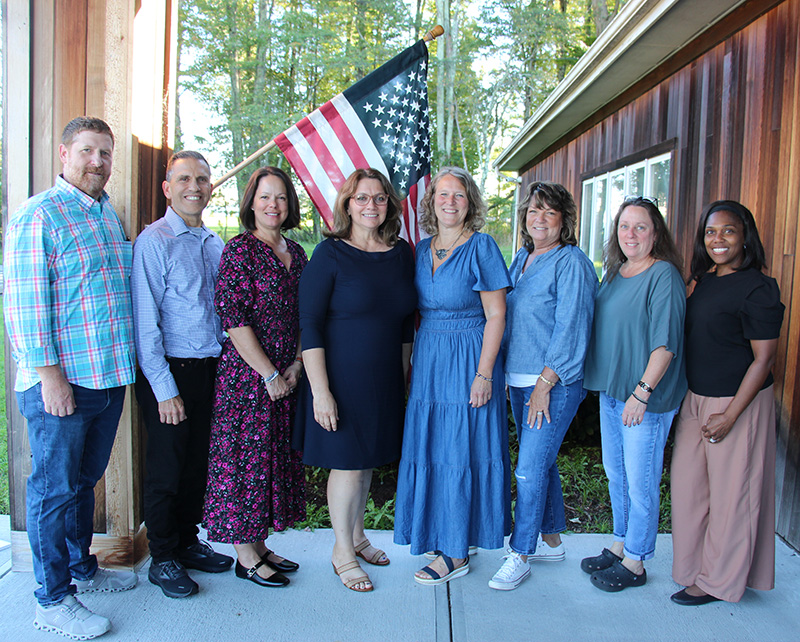 A group of eight adults, two men on right followed by six women. There is an American flag behind them.