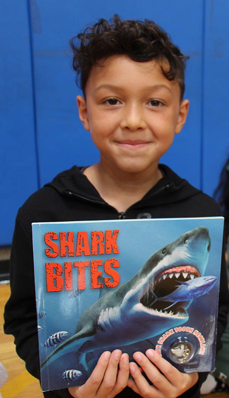 A boy holds up a book that says Shark Bites. He is smiling.