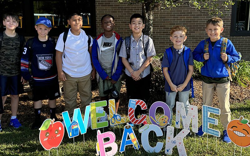 Seven elementary age boys smile as they stand under a tree and behind letters that say Welcome Back.