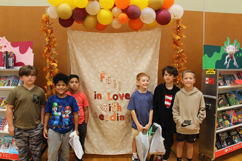 A group of six third-grade boys stand on either side of a large sign that says Fall in love with reading. There are yellow, orange, white and brown balloons along the top.