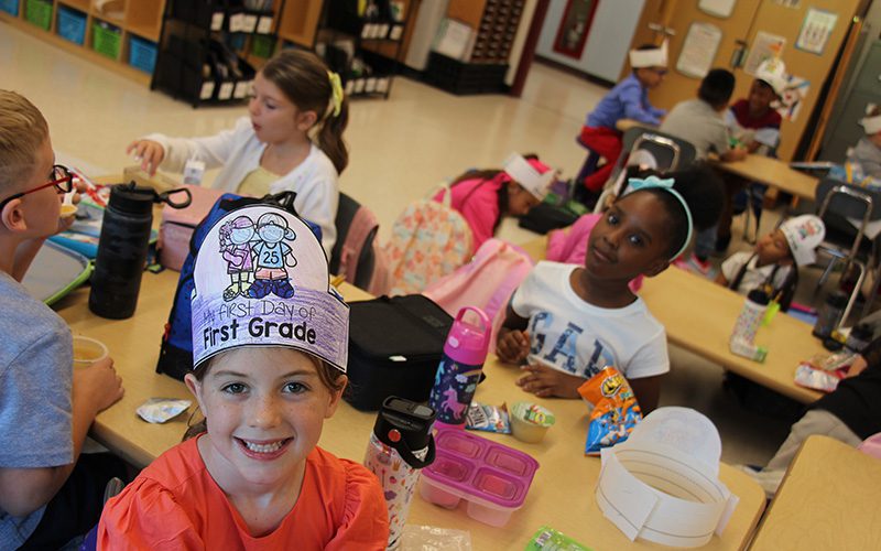 A young elementary student smiles wearing a paper hat that says First day of first grade. Behind her is a table with several students at it.