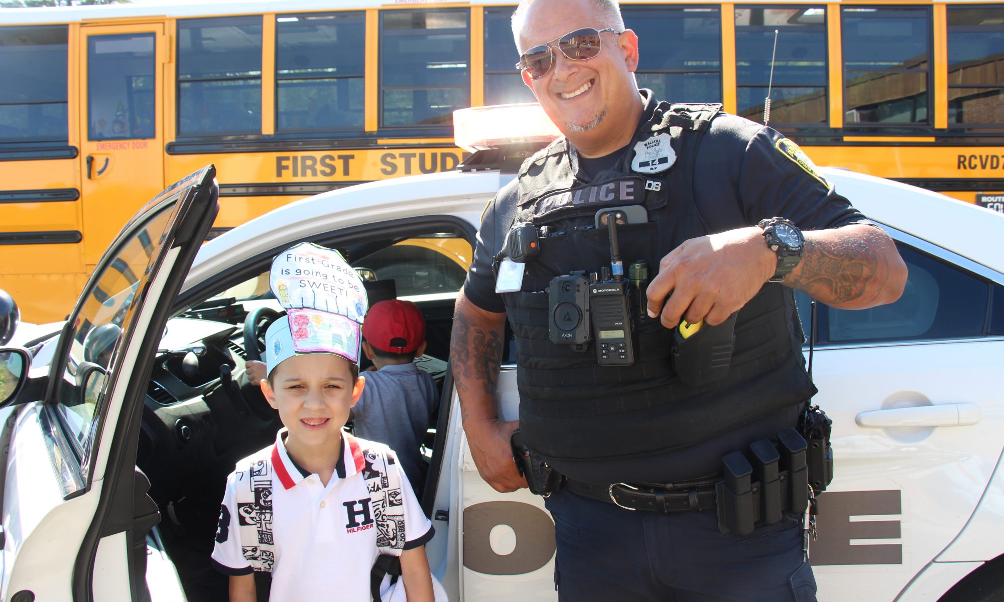 With a yellow school bus in the background, a kid wearing a paper hat that says First grade s going to be sweet stands with a police officer. Behind them is a police car with a kid sitting in it.