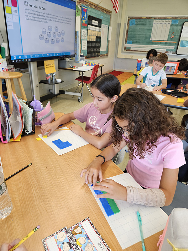 Two fourth-grade girls work at desks creating a table with small multi-colored tiles.