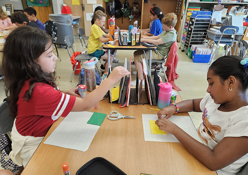 Two fourth-grade girls sit at desks using multi-colored tiles to create a graph.