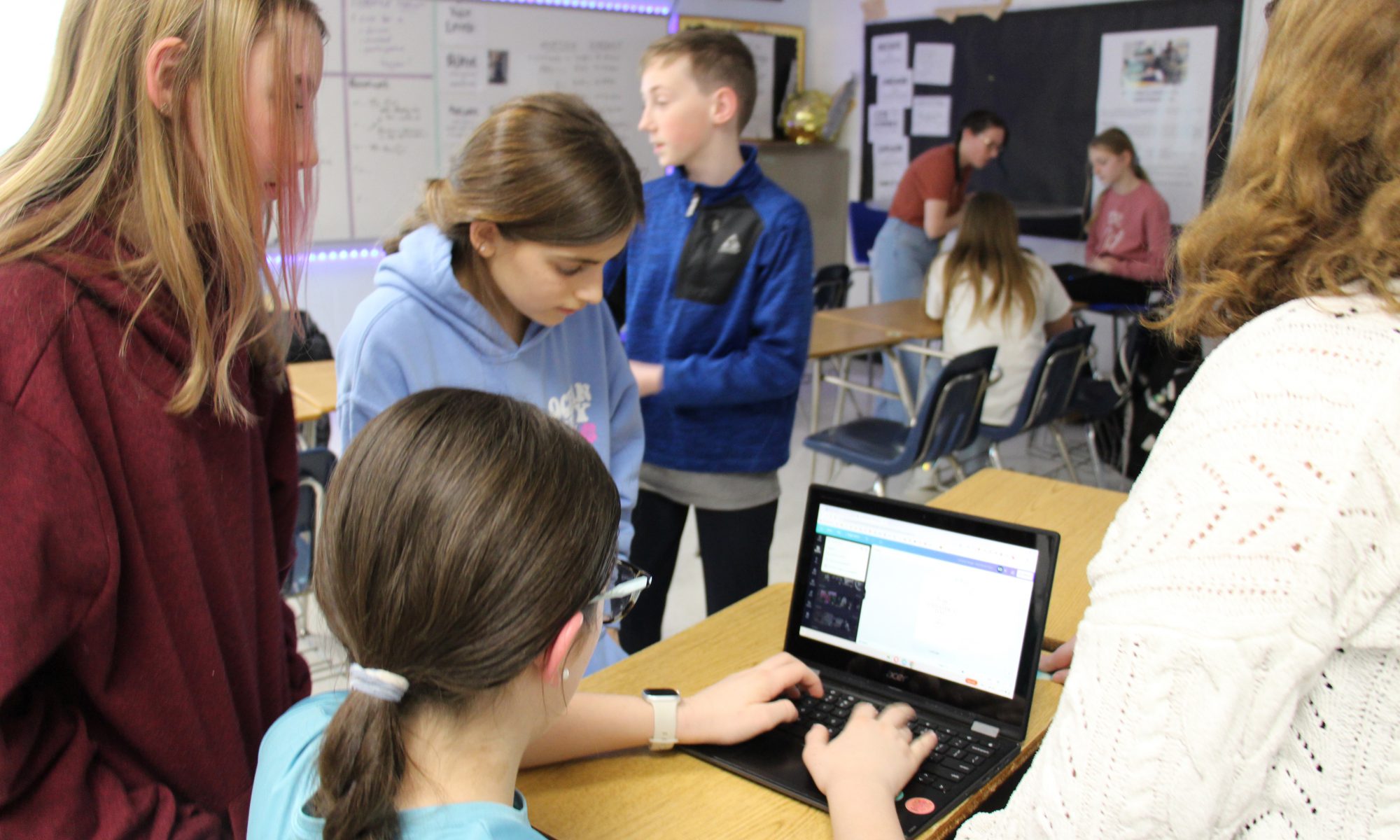 A middle school girl sits working on a Chromebook while three other girls and one boy stand around her. They are all working on a flyer.