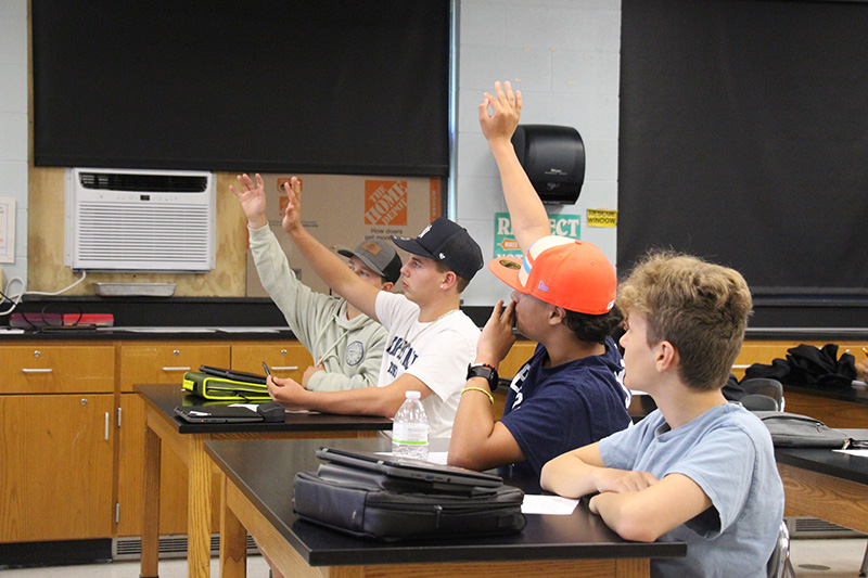 Four middle school boys sitting at lab tables raise their hands.