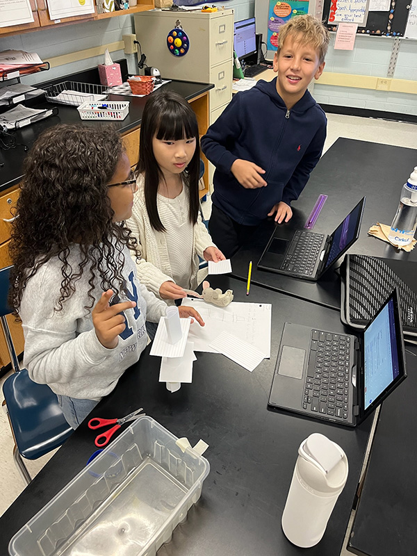 Three sixth-grade students - two girls and a boy - work on building a tower made of index cards.