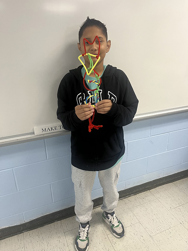 A sixth grade kid smiles as he holds up a structure he built with pipe cleaners.