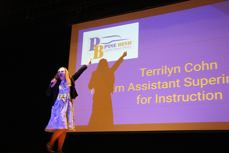 A woman in a dress with long blonde hair holds a microphone an dpoints up at a screen. the screen has a PB logo and the words Terrilyn Cohn Interim Assistant Superintendent for Instruction