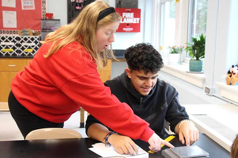 A woman with long blonde hair, wearing a red sweater points out something on a scale to a young man with dark hair wearing a black sweatshirt.