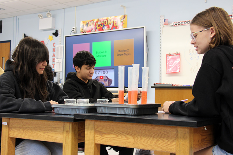 Eighth-grade students sit at lab tables with beakers of liquids in front of them.