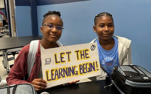 Two middle school kids sit at a table and hold a sign that says Let the Learning Begin! They are both smiling.