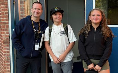 A man on the left smiles as he holds open a school building door. There are two middle school kids standing with him smiling.