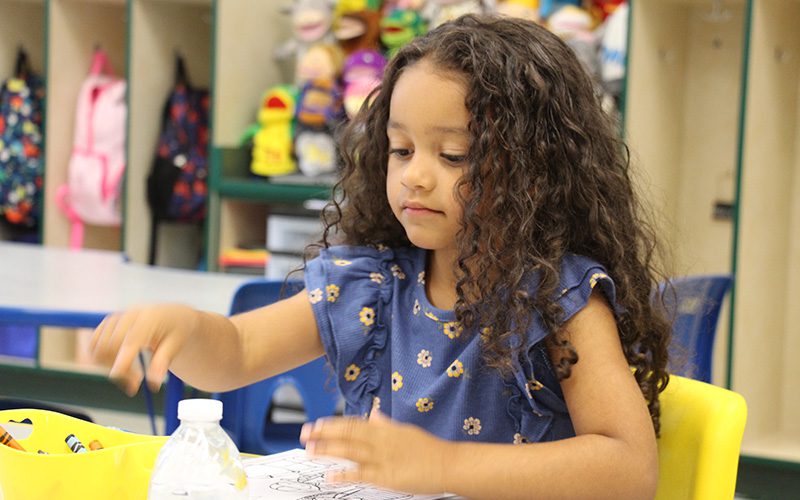 A younger elementary student with long dark hair, wearing a blue and white print shirt, reaches her hand for a crayon.