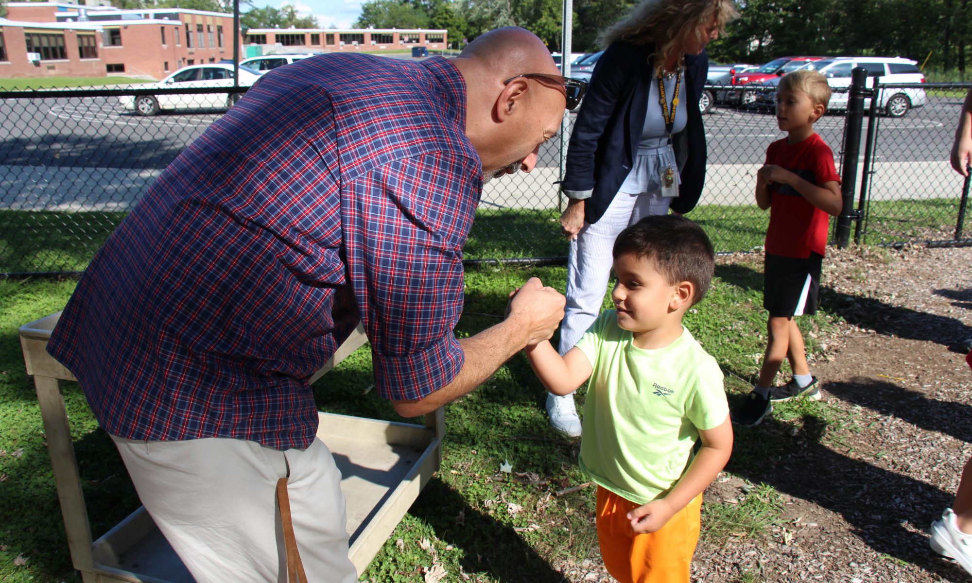 A man in a blue and red plaid shirt with sunglasses on top of his head gives a fistbump to a small child. The kid has short dark hair and is wearing orange shorts and a light green shirt.