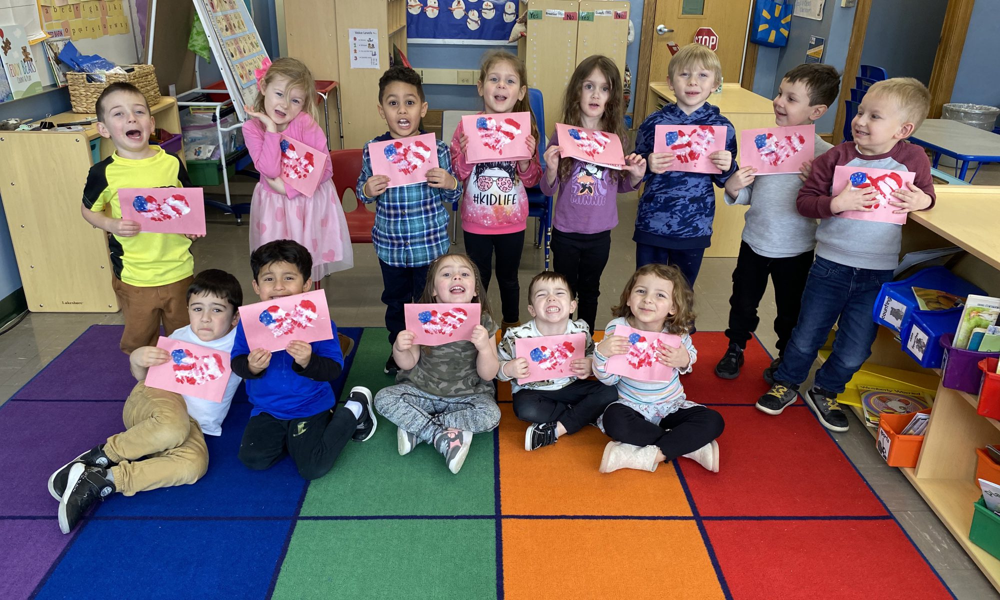 Thirteen pre-K students standing in two rows on a multi-colored rug. They are holding pink valentines painted red, white and blue they made for veterans.