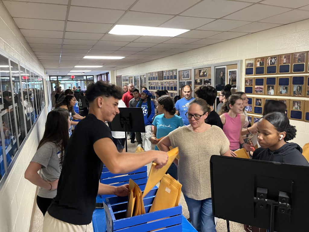 A large group of people in a hallway. High school kids are behind a table handing out large envelopes to the people on the other side of the table.