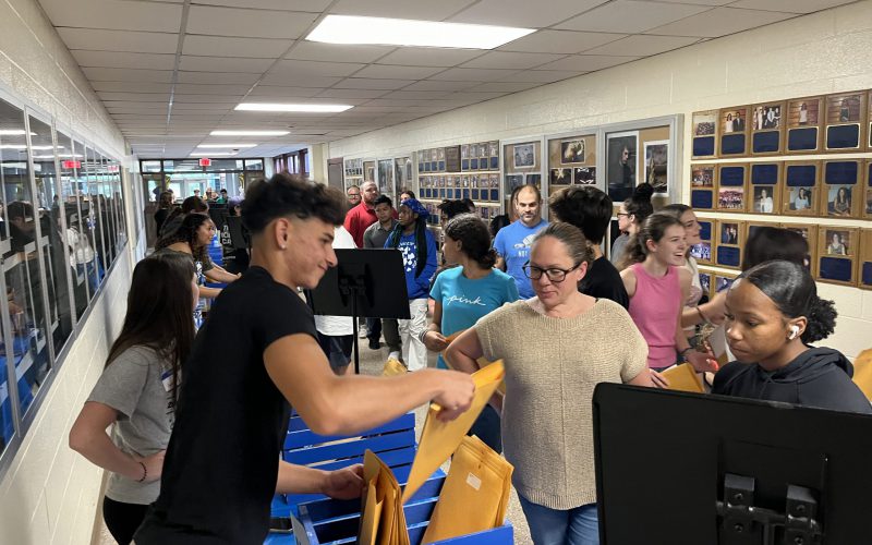 A large group of people in a hallway. High school kids are behind a table handing out large envelopes to the people on the other side of the table.