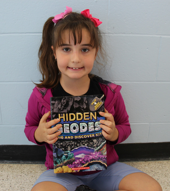 A girl with dark hair smiles as she holds up a box that says Hidden Geodes.