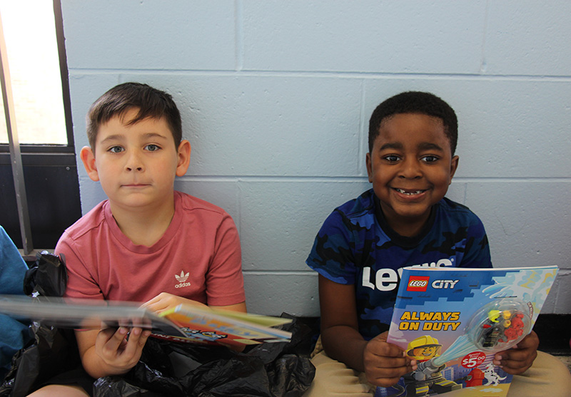 Two first-grade boys sit on a floor holding up the books they just bought.