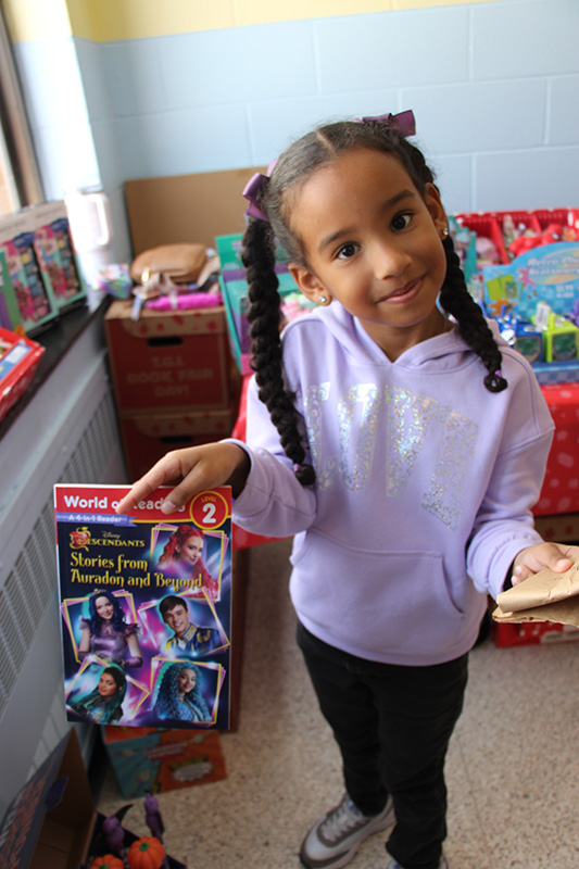 A little girl with long braids smiles as she holds up her book. she is wearing a lavender shirt.