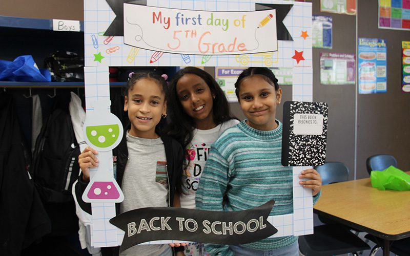 Three fifth-grade girls smile and hold a frame around them that says My first day of 5th grade.