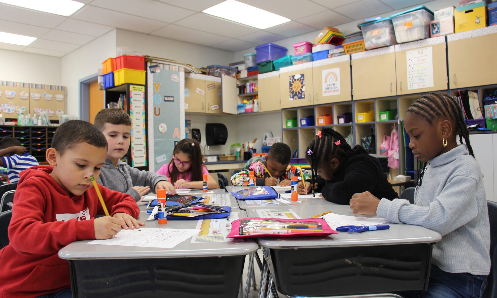 A group of six second graders work at desks that are pushed together .