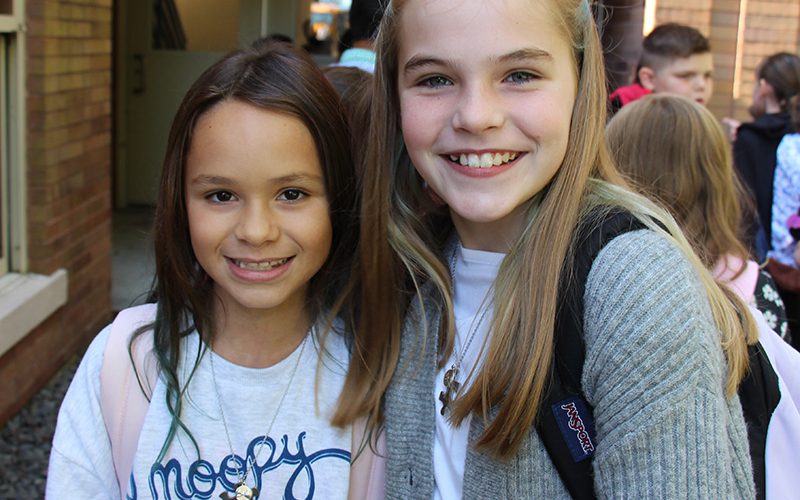 Two elementary school girls, one with long dark hair and one with long blonde hair, smile.