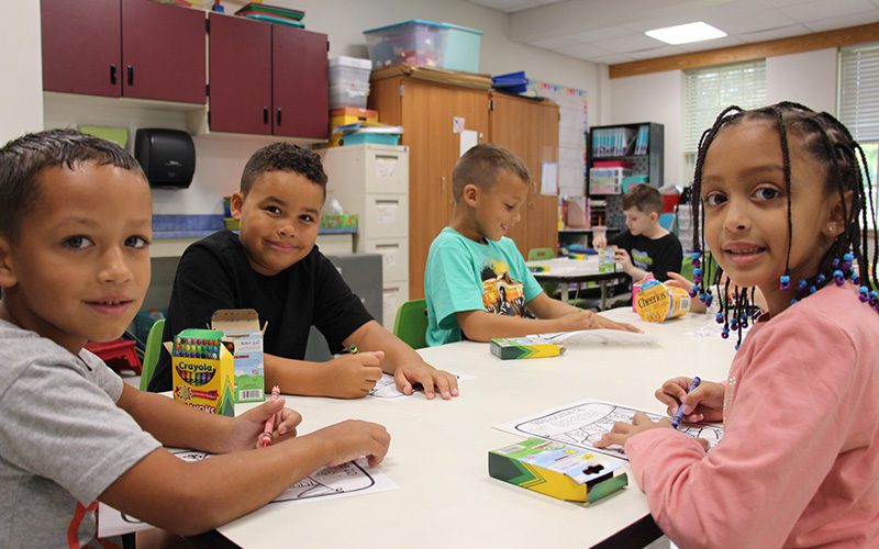 A group of four elementary students sit at a table and color. They are looking at the camera and smiling.