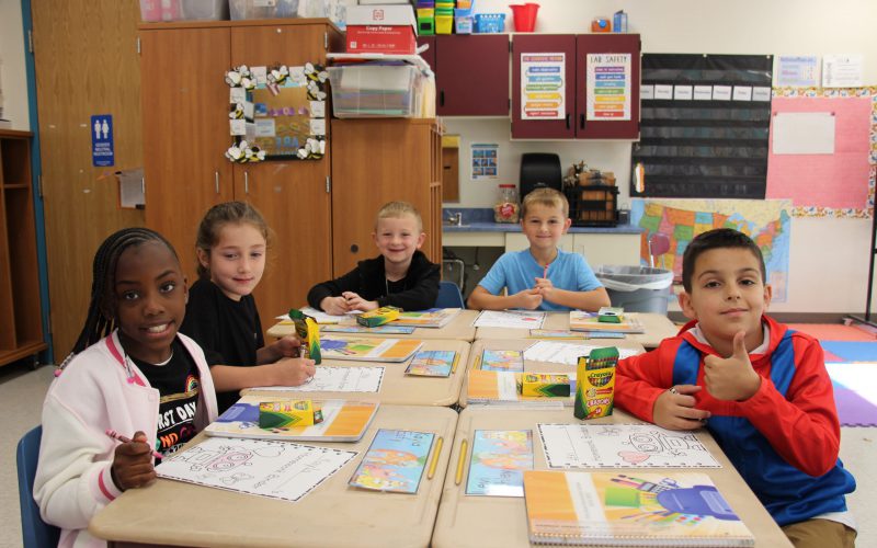 A group of younger elementary students sit at desks pushed together