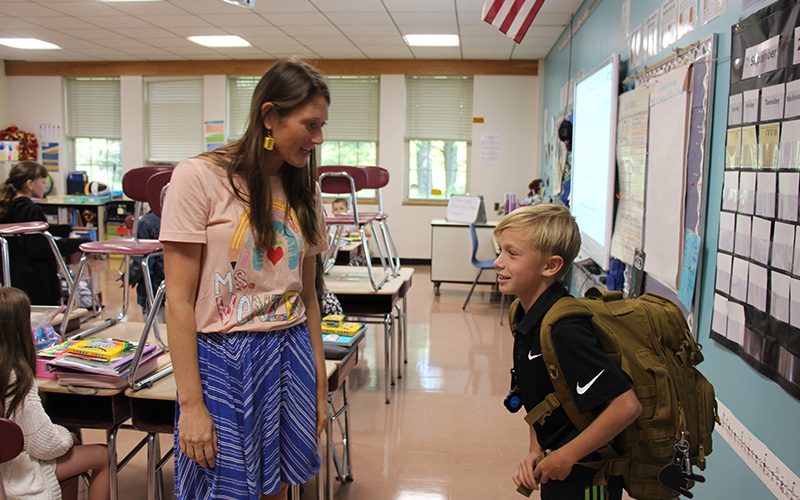 A woman with long dark hair talks to an elementary student, a boy with short blonde hair, carrying a large backpack.