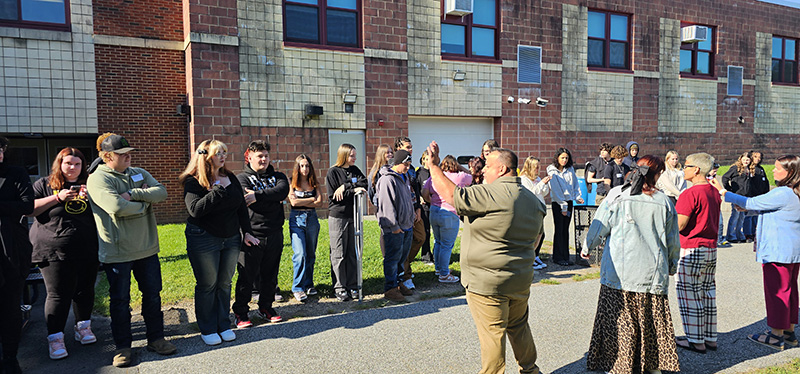 A line of high school students stand and listen to a man standing in front of them.