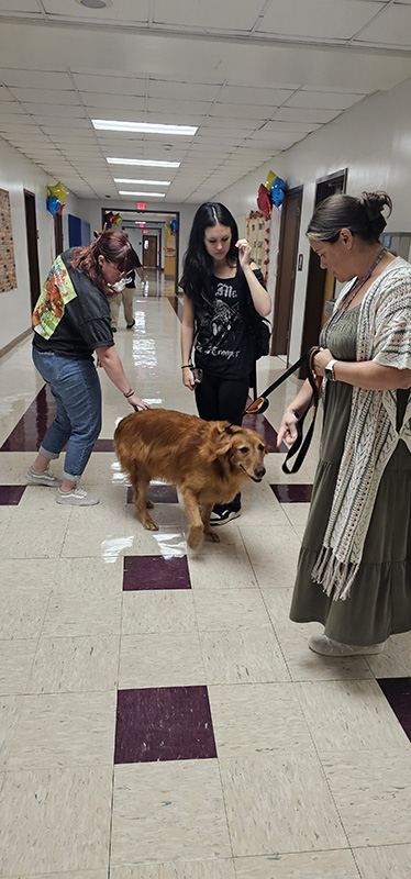 A large golden retriever stands in a group of adults and kids with a high school student petting him.