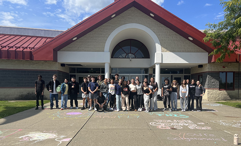 A large building with a red roof and a group of 29 students standing in front. There are positive messages written in chalk on the sidewalk in front of them.