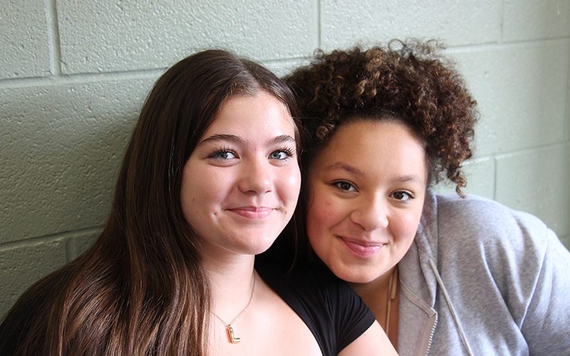 Two girls sit with their heads close together smiling. the girl on the left has long dark hair and the girl on the right has curly dark hair.