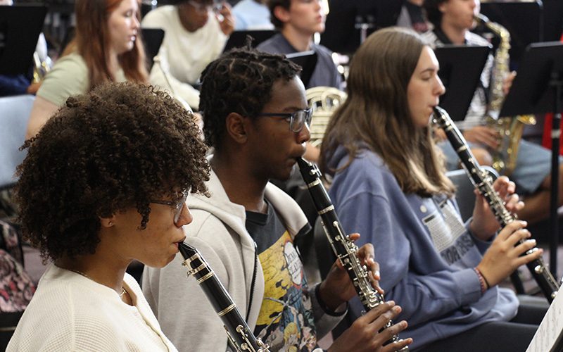 Three high school kids play clarinets in a line. Behind them are other musicians.