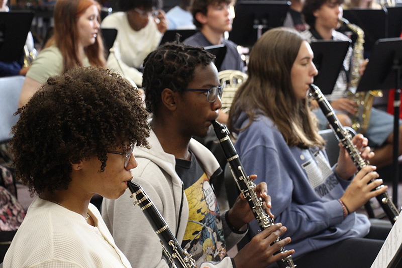 Three high school kids play clarinets in a line. Behind them are other musicians.