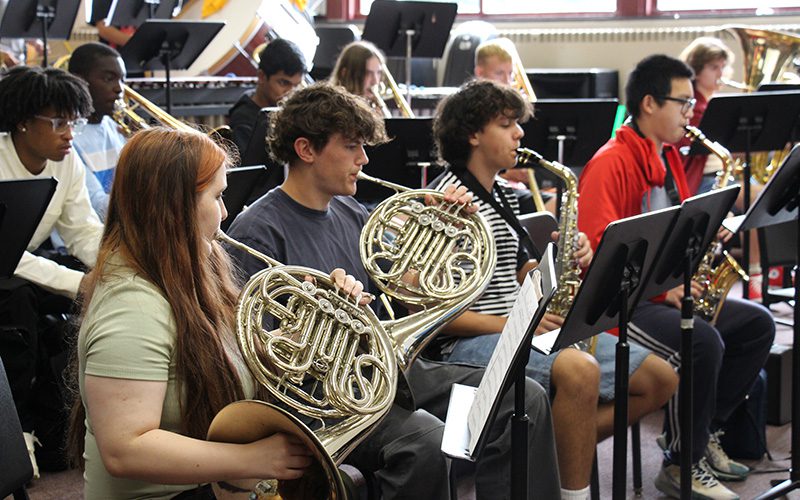 Four high school musicians sit and play their instruments in a row. Two are French horn and two are saxophones.