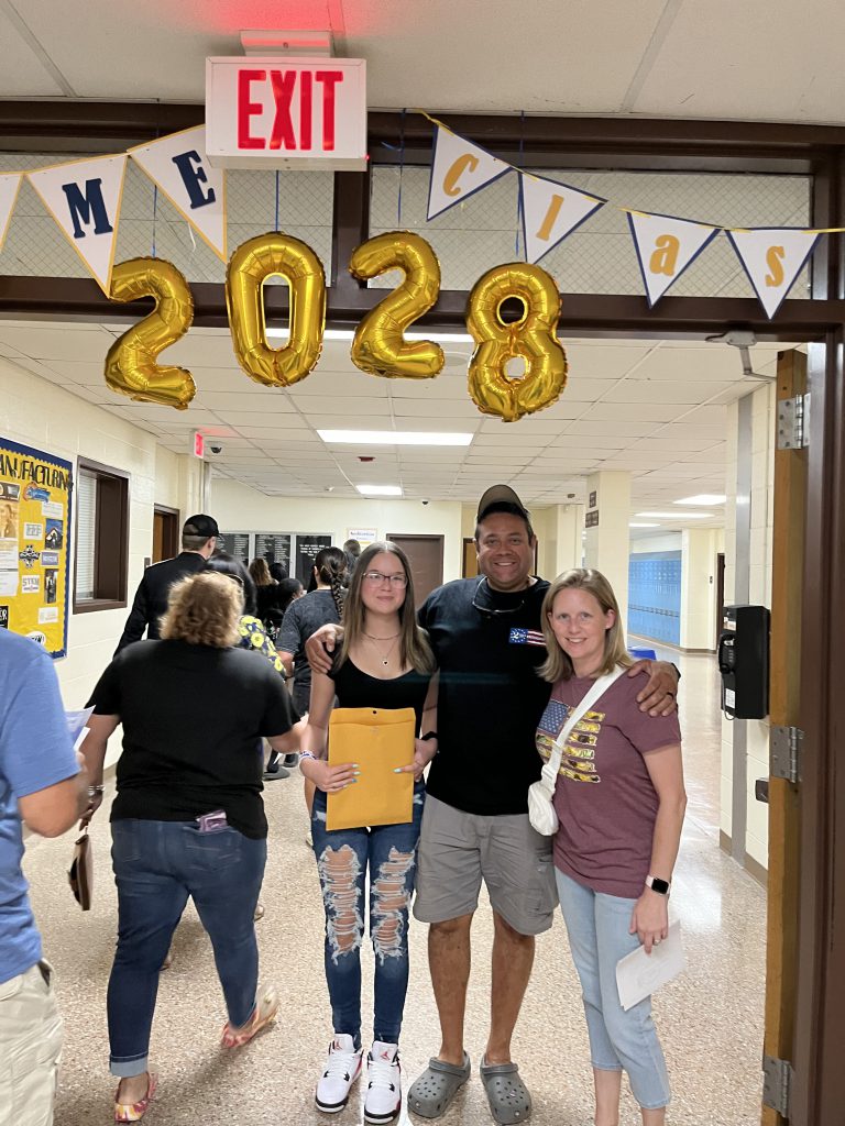 Large gold balloons say 2028 and under them are a high school student with a man an a woman.