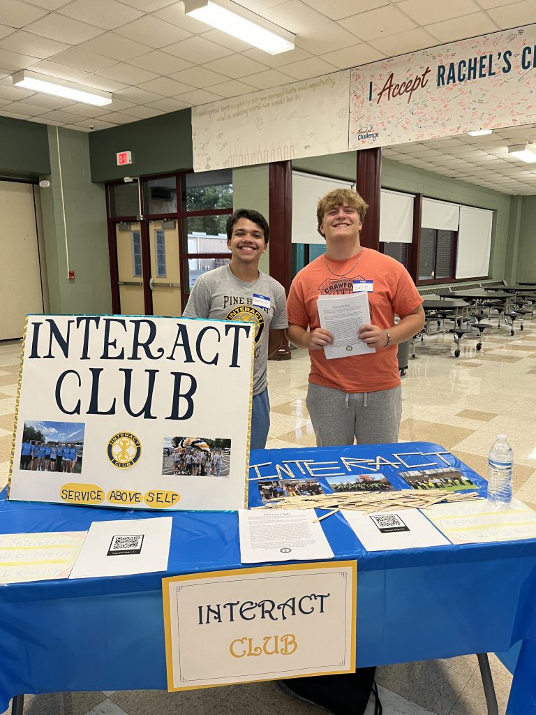 Two high school boys stand behind a table with a large sign that says Interact Club