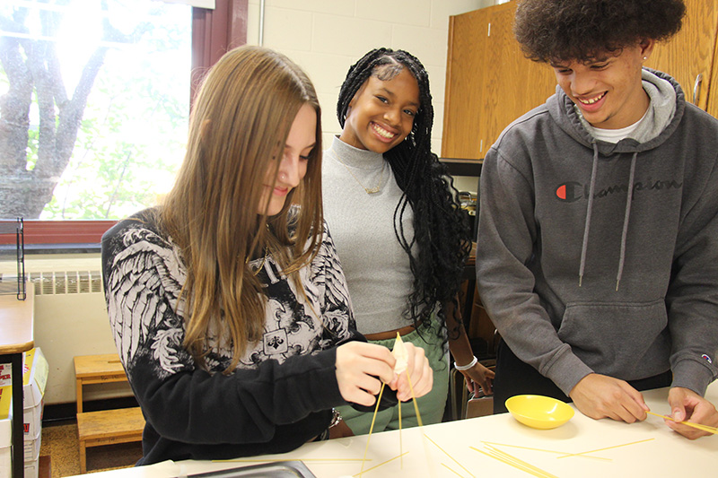 Three high school students are at a table building a tower made from spaghetti.