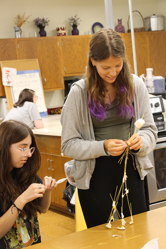 Two high school girls work together on a tower made of spaghetti.