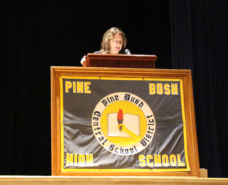 A woman with medium length dark hair stands at a podium speaking. In front of the podium is a large framed banner that says Pine Bush High School with a circle in the center.