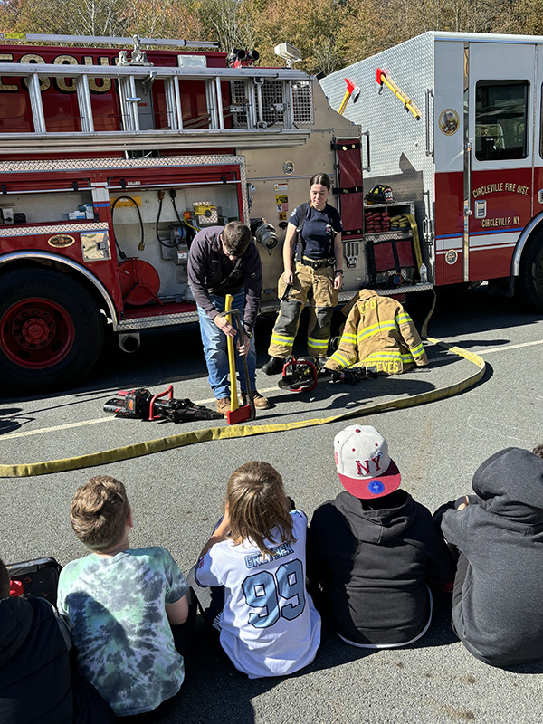 A group of elementary students sit and listen as fire fighters show equipment from their fire truck. There is a red fire truck in the back ground.