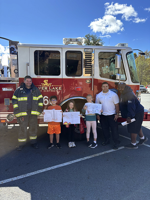 Three elementary students stand holding their posters. They are in front of a red and white fire truck. Next to them is a fire fighter on either side. On the right there are two adults. 